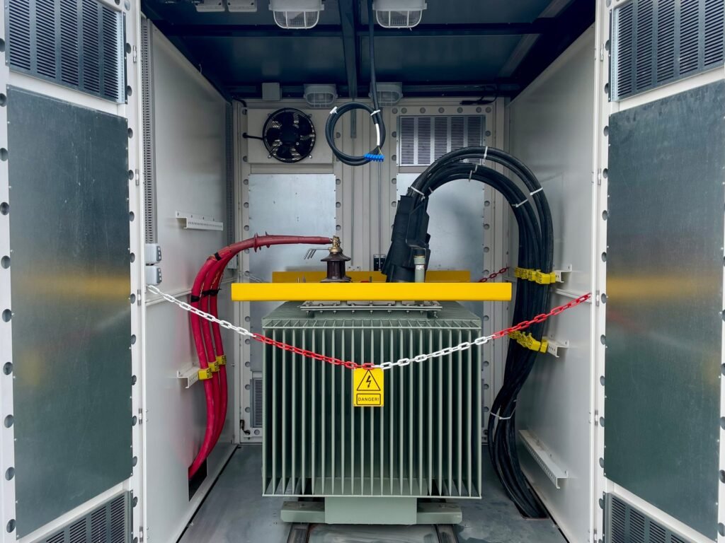 Oil-filled distribution transformer in the transformer compartment of a compact substation metal kiosk, with medium-voltage and low-voltage cable connections and a safety chain across the opening